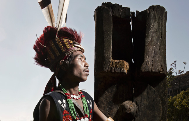 Pangmi Naga warrior in front of a pole with carved gong, symbol of wealth.