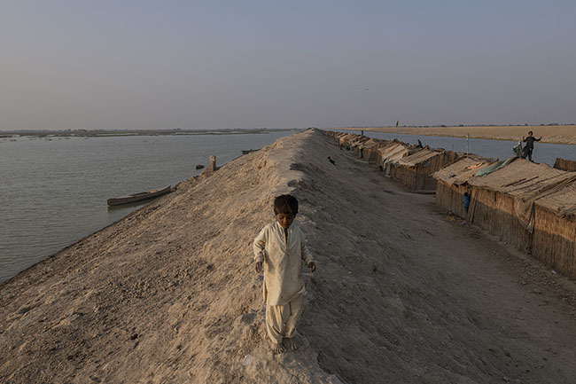 A Mohana child plays on the raised banks of the Right Bank Outfall Drain (RBOD), where huts made of tarps, reeds, and scavenged materials line up.