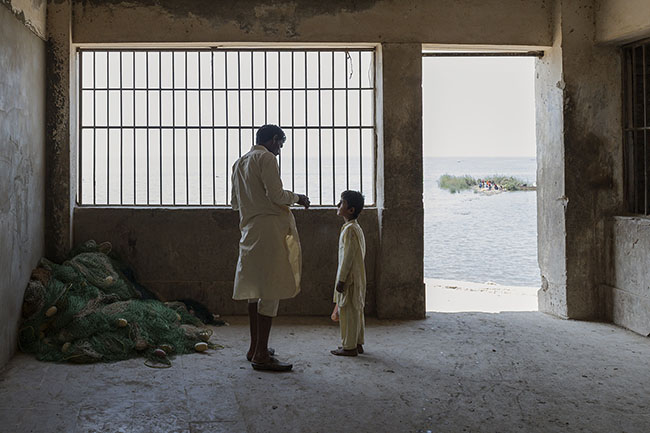 A father speaks to his son behind the bars of the only fish market on Lake Manchar. Once, the fishing of the Mohana fed an entire region. Today, it no longer sustains families. And with it, an entire culture teeters, drifting on a lake that is but a mirage of what it once was. But the Mohana still cling to the hope of a better future for their children.