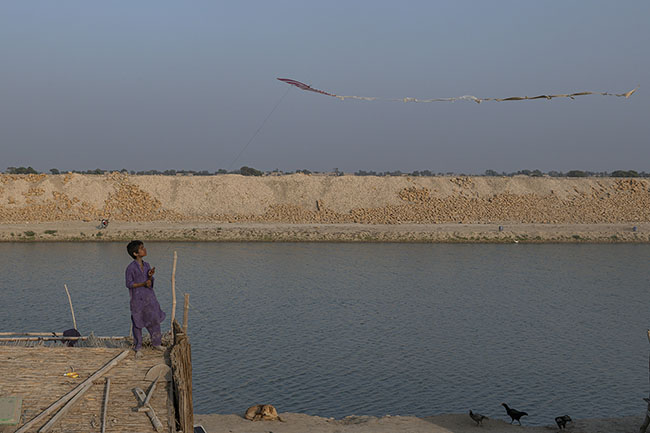 At the edge of the Right Bank Outfall Drain (RBOD), a Mohana child flies his kite from the roof of a hut. Despite the difficulties, the gestures of childhood remain: light and fleeting like the wind.