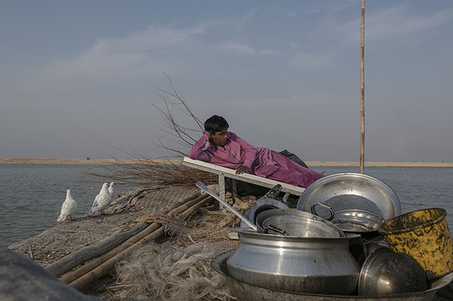 A Mohana child rests on the roof of a boat. Around him, dry wood, washing utensils, a fishing net, and a solar panel pile up, providing them with some electricity.