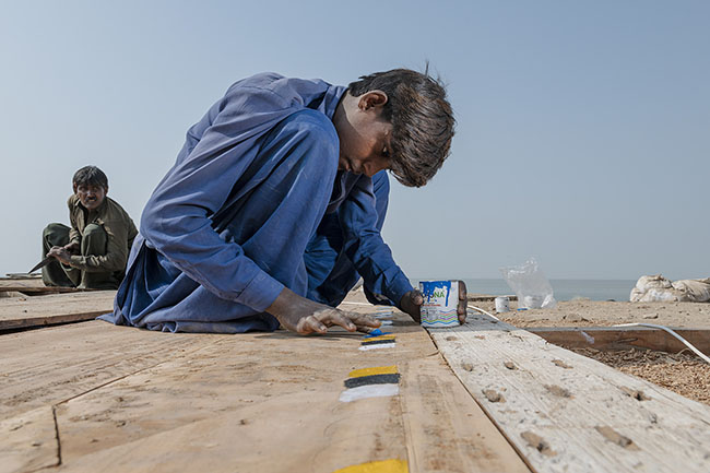 A Mohana child hand-paints traditional motifs on a board that will be used in the construction of a boat.